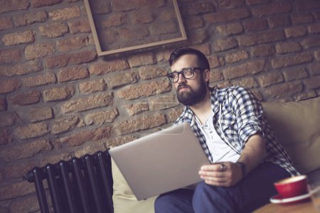 Photo for Handsome hipster guy sitting on a sofa in a cafe, having a morning coffee and using a laptop computer for his freelancing job - Royalty Free Image