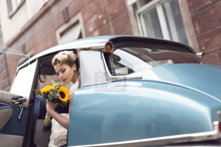 Téléchargez les photos : Belle jeune mariée assise dans une robe de mariée dans une vieille voiture rétro, tenant un bouquet de tournesol - en image libre de droit