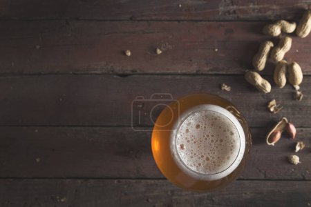 Téléchargez les photos : Vue de dessus d'un verre de bière pâle non filtrée avec de la mousse et quelques cacahuètes sur une table de pub rustique en bois - en image libre de droit