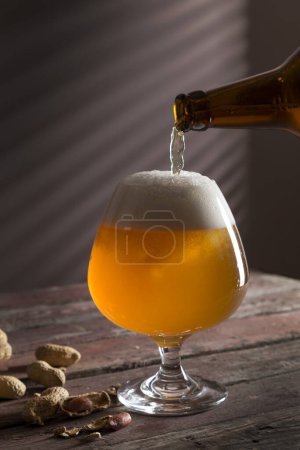 Foto de Detalle de un vaso de cerveza pálida fría sin filtrar que se vierte de una botella, con algunos cacahuetes al lado del vaso. Enfoque selectivo - Imagen libre de derechos