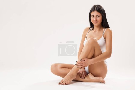 Téléchargez les photos : Belle jeune femme portant des sous-vêtements en coton blanc, assise isolée sur fond blanc, regardant vers la caméra - en image libre de droit