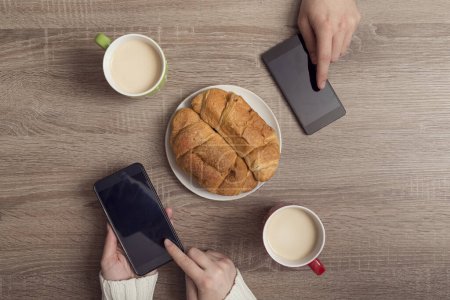 Foto de Vista superior de manos masculinas y femeninas sosteniendo teléfonos inteligentes y navegando por la red mientras toma café y desayuna - Imagen libre de derechos