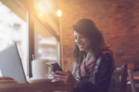 Téléchargez les photos : Jeune femme moderne assise dans un café, prenant un café le matin, travaillant sur un ordinateur portable et envoyant un message texte sur son téléphone intelligent. - en image libre de droit