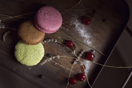 Photo for Top view of macaron cookies on a wooden tray, with cherry fruit and lavender flowers placed next to it. Selective focus on brown and pink cookie - Royalty Free Image