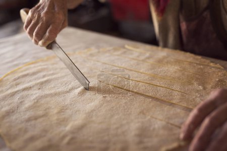 Téléchargez les photos : Gros plan de la main d'une femme âgée tenant un vieux couteau et coupant des lanières de pâte fine roulée pour faire des pâtes maison. Concentration sélective - en image libre de droit