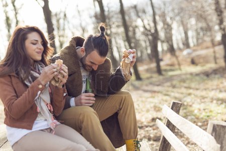 Téléchargez les photos : Jeune couple assis sur un banc en bois au bord de la route forestière, profitant d'une journée ensoleillée d'automne, mangeant des sandwichs et buvant de la bière. Concentrez-vous sur le gars - en image libre de droit