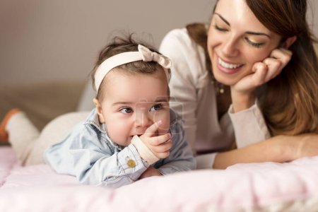 Photo for Beautiful baby girl and her mother lying in bed, playing and cuddling. Baby chewing her hands. Focus on the baby - Royalty Free Image