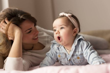 Photo for Beautiful baby girl and her mother lying in bed, playing and cuddling. Focus on the baby - Royalty Free Image