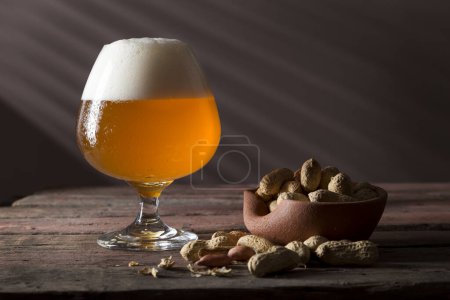 Foto de Un vaso de cerveza fría sin filtrar y un tazón de maní en una mesa rústica de pub de madera. Concéntrate en la espuma - Imagen libre de derechos