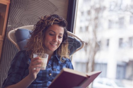 Téléchargez les photos : Belle jeune femme assise dans un café à côté d'une fenêtre, pensant et lisant un livre - en image libre de droit