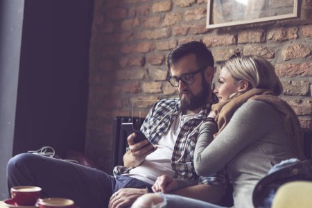 Téléchargez les photos : Jeune couple assis dans un café, prendre un café le matin, surfer sur le web sur un téléphone intelligent et profiter de leur temps ensemble. Concentrez-vous sur le gars - en image libre de droit