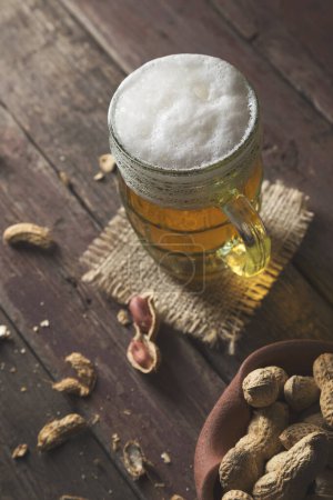 Photo for Mug of light beer with froth placed on a burlap cover and some peanuts on a rustic wooden pub table. Focus on the froth - Royalty Free Image