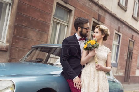 Téléchargez les photos : Couple nouvellement marié debout dans la rue pavée à côté d'une vieille voiture rétro, embrasser et embarquer sur une lune de miel - en image libre de droit