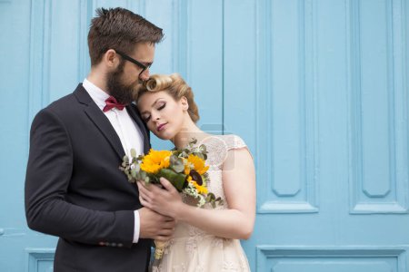 Téléchargez les photos : Couple nouvellement marié debout à côté d'une porte en bois rétro bleu, tenant un bouquet de mariage de tournesol et câlin - en image libre de droit