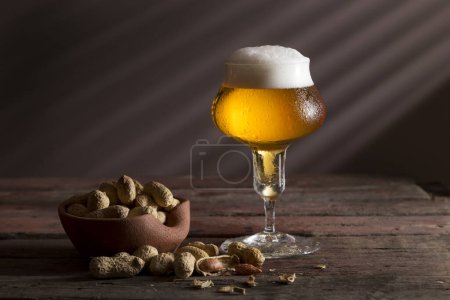 Foto de Vaso de cerveza fría pálida con un tazón de maní sobre una mesa rústica de madera - Imagen libre de derechos