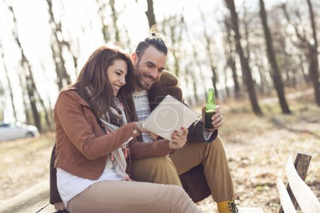 Photo for Couple in love sitting on a wooden bench in the forest on a beautiful autumn day, holding a tablet computer and searching for hiking tracks. Focus on the guy - Royalty Free Image