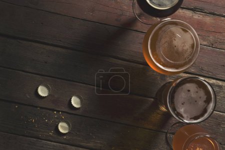 Téléchargez les photos : Vue de dessus d'une bière pâle, foncée, non filtrée et aux fruits rouges dans quatre verres à bière différents sur une table en bois rustique. Focus sur la table et les bouchons de bière - en image libre de droit