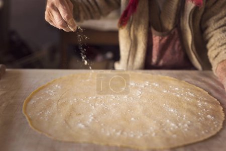 Téléchargez les photos : Détail de la main d'une femme âgée saupoudrer de farine sur une fine pâte roulée tout en faisant des pâtes maison. Focus sélectif sur une pâte - en image libre de droit