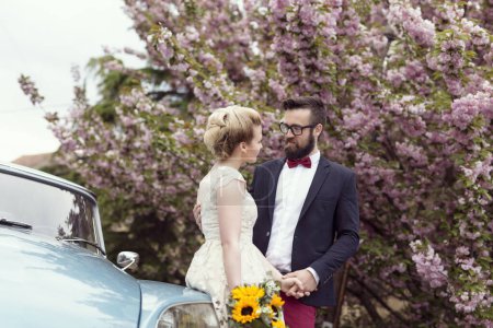 Photo for Newlywed couple standing in the street next to an old retro car, hugging and embarking on a honeymoon - Royalty Free Image