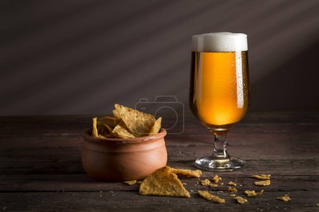 Foto de Vaso de cerveza fría pálida con un tazón de patatas fritas sobre una mesa rústica de madera - Imagen libre de derechos