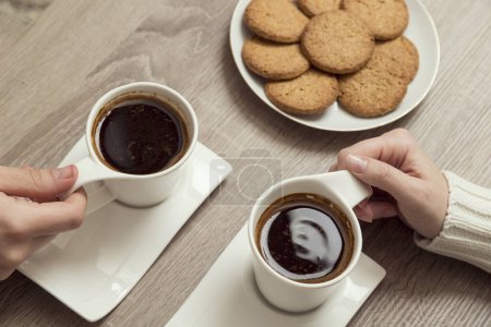 Photo for Top view of male and female hands holding cups of american coffee with plate of chocolate chip cookies placed next to it. Focus on the female's hands and cup of coffee. - Royalty Free Image