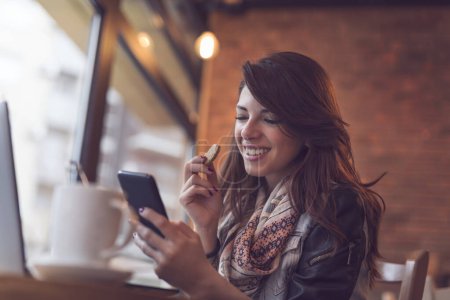 Foto de Mujer joven y moderna sentada en un café, tomando café por la mañana, trabajando en una computadora portátil y enviando mensajes de texto en su teléfono inteligente. - Imagen libre de derechos