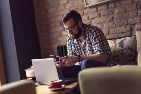 Téléchargez les photos : Beau mec hipster assis dans un fauteuil dans un café, prenant un café du matin et utilisant un ordinateur portable et un téléphone intelligent pour son travail indépendant - en image libre de droit