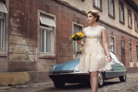Photo for Beautiful young bride posing in a wedding dress in a retro cobble street, with an old timer car in the background, holding a sunflower bouquet - Royalty Free Image