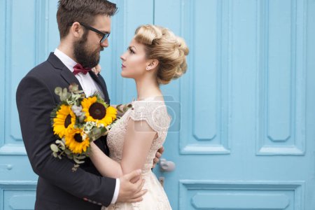 Téléchargez les photos : Couple nouvellement marié debout à côté d'une porte en bois rétro bleu, tenant un bouquet de mariage de tournesol et se regardant - en image libre de droit