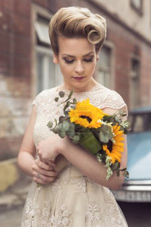Téléchargez les photos : Belle jeune mariée posant dans une robe de mariée dans une rue pavée rétro, avec une vieille voiture de minuterie en arrière-plan, tenant un bouquet de tournesol - en image libre de droit