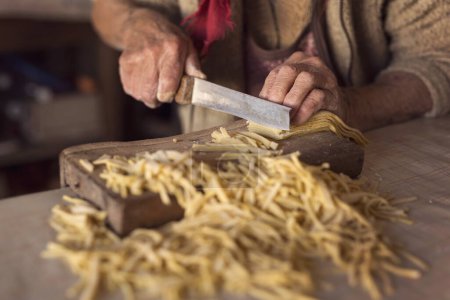 Photo for Detail of an elderly woman's hand cutting noodles with an old knife while making homemade pasta. Selective focus - Royalty Free Image