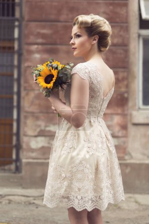 Téléchargez les photos : Belle jeune mariée posant dans une robe de mariée dans une rue pavée rétro, tenant un bouquet de tournesol - en image libre de droit