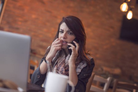 Téléchargez les photos : Jolie jeune brune assise dans un café et ayant une conversation téléphonique - en image libre de droit