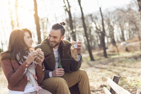 Téléchargez les photos : Jeune couple assis sur un banc en bois au bord de la route forestière, profitant d'une belle journée de printemps et mangeant des sandwichs - en image libre de droit