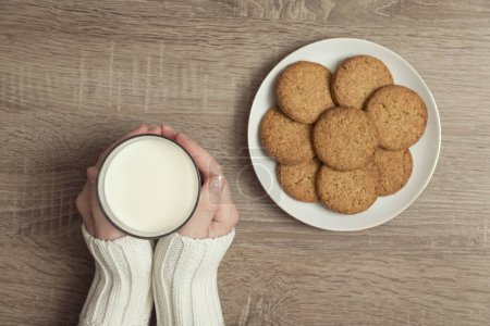 Téléchargez les photos : Vue de dessus des mains féminines tenant un verre de lait, avec une plaque de biscuits aux pépites de chocolat placée à côté sur la table. Focus sur le pouce - en image libre de droit