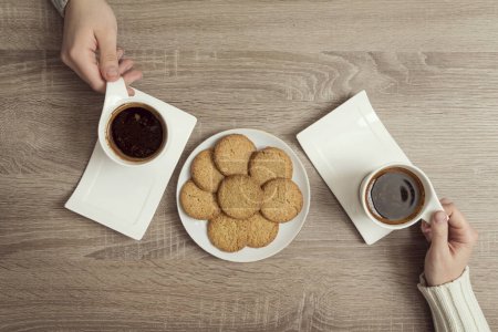 Photo for Top view of male and female hands holding cups of american coffee with plate of chocolate chip cookies placed next to it - Royalty Free Image