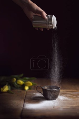 Téléchargez les photos : Détail d'une main féminine tenant une sucrerie et saupoudrant de sucre en poudre sur une tasse à biscuits. Concentration sélective - en image libre de droit