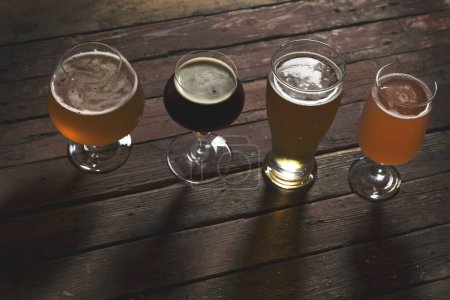 Foto de Cerveza pálida, oscura, sin filtrar y de fruta roja en cuatro vasos de cerveza diferentes sobre una mesa de madera rústica. Enfoque selectivo - Imagen libre de derechos