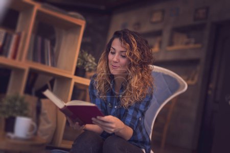 Téléchargez les photos : Belle jeune femme assise dans un café, profitant de son café du matin et lisant un livre - en image libre de droit