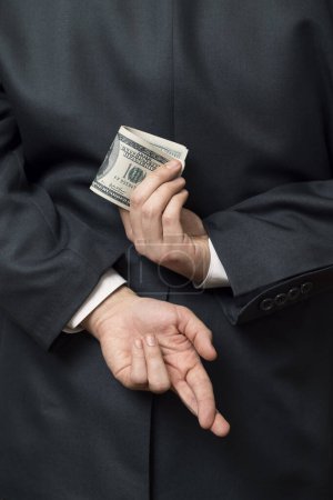 Téléchargez les photos : Détail d'un homme d'affaires debout et tenant ses mains derrière son dos, tenant un tas d'argent dans une main et les doigts croisés sur un autre. Concentration sélective - en image libre de droit
