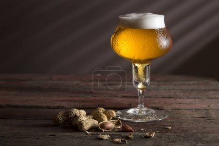 Foto de Vaso de cerveza fría pálida con un tazón de maní sobre una mesa rústica de madera. Enfoque selectivo - Imagen libre de derechos