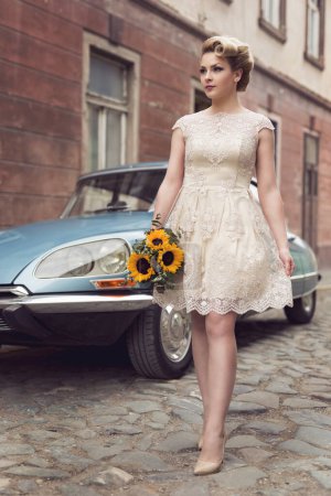 Téléchargez les photos : Belle jeune mariée posant dans une robe de mariée dans une rue pavée rétro, avec une vieille voiture de minuterie en arrière-plan, tenant un bouquet de tournesol - en image libre de droit