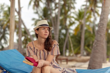 Téléchargez les photos : Jeune femme assise sur un transat sous les palmiers dans une magnifique station balnéaire exotique d'île tropicale, lisant un livre et se relaxant tout en profitant du lever du soleil sur la mer - en image libre de droit