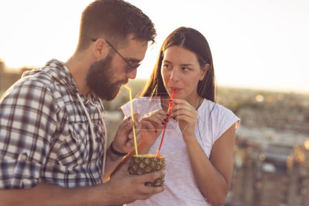 Photo for Young couple in love drinking a pineapple cocktail and having fun at rooftop party in sunset - Royalty Free Image