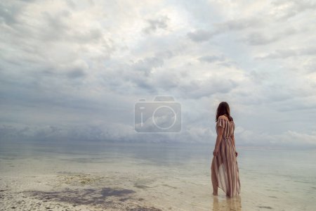 Téléchargez les photos : Belle jeune femme marchant sur la plage isolée à l'aube, profitant d'un beau lever de soleil sur l'océan Indien ; femme se relaxant à la plage profitant du coucher de soleil sur la mer - en image libre de droit