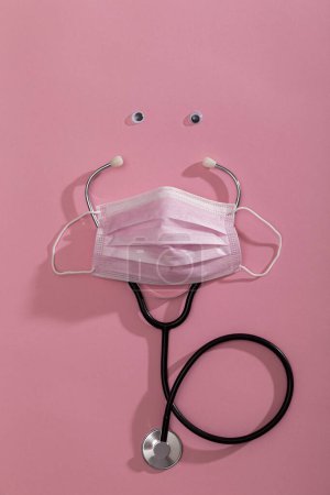 Foto de Vista superior de la máscara de protección facial quirúrgica rosa, estetoscopio y ojos pequeños aislados en el fondo de color rosa, como símbolo de especialistas en atención médica que luchan contra la pandemia covid-19 - Imagen libre de derechos