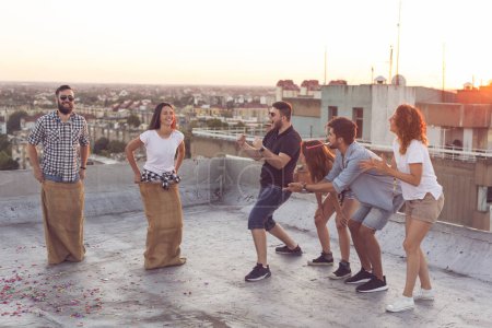 Téléchargez les photos : Groupe de jeunes gens qui s'amusent à la fête d'été sur le toit, acclamant leurs concurrents dans une course de sac - en image libre de droit