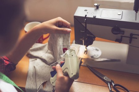 Téléchargez les photos : Couture attachant une étiquette sur un T-shirt nouvellement fabriqué avec un pistolet d'étiquette. Focus sélectif sur le bouton rouge - en image libre de droit