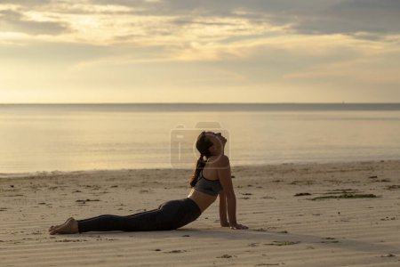 Foto de Mujer joven ejercitándose en la playa temprano en la mañana, practicando yoga, haciendo ejercicios de estiramiento, sosteniendo hacia arriba mirando hacia atrás al perro doblando la pose de yoga - Imagen libre de derechos