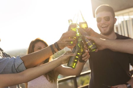Foto de Grupo de jóvenes amigos divirtiéndose en la fiesta en la azotea, bebiendo cerveza, haciendo un brindis y disfrutando de los calurosos días de verano. Enfoque selectivo en las botellas de cerveza y las manos - Imagen libre de derechos